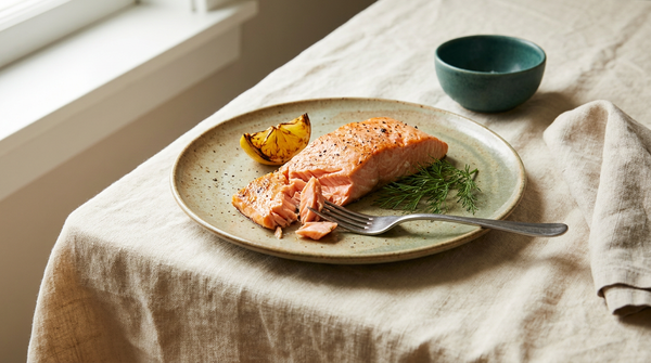 A cooked salmon fillet on a ceramic plate with lemon and dill, on cream linen in soft daylight