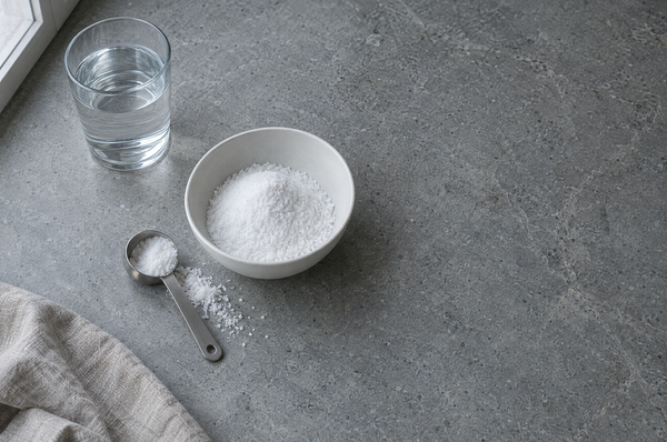 A white ceramic bowl of fine white powder beside a stainless steel measuring scoop and a glass of water on a grey stone kitchen worktop.